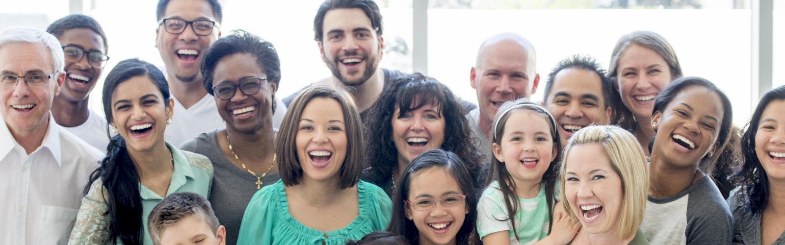 A diverse group of people, including men, women, and children, smiling and standing close together indoors.