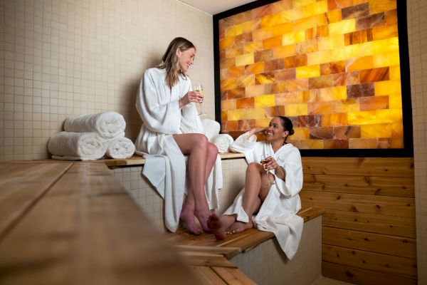 Two women in bathrobes relax and chat while drinking in a sauna room, with folded towels in the background and a warm, illuminated wall.