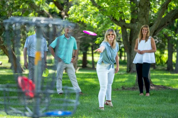 Four people are playing disc golf in a park, with one woman throwing a disc towards the basket while the others watch.