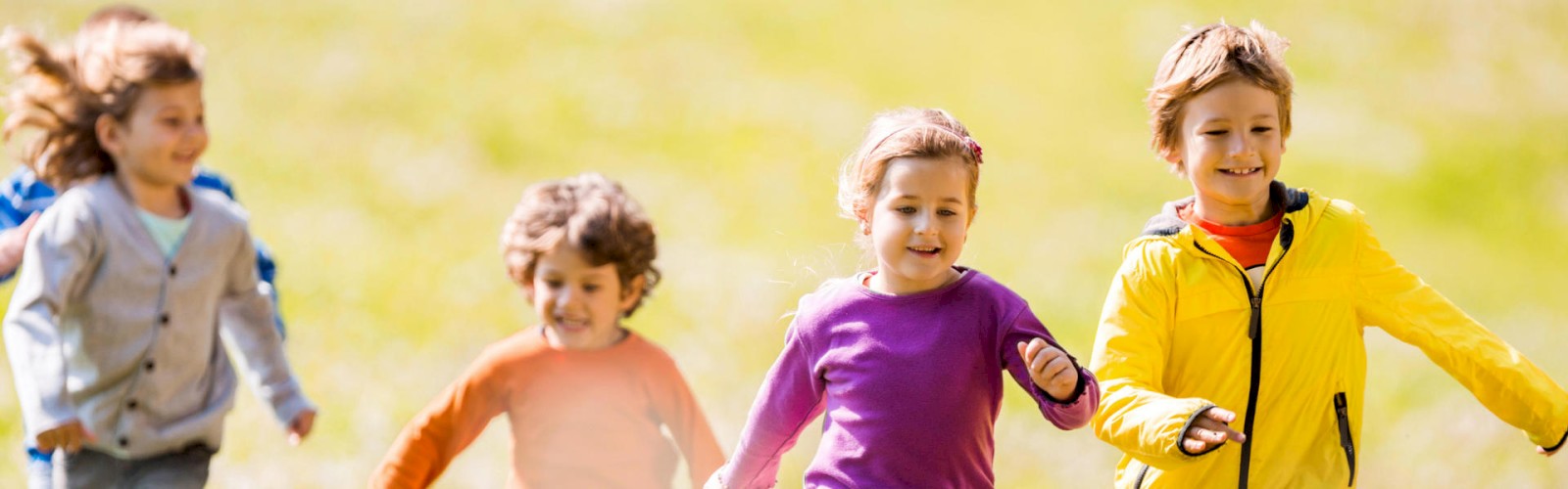 The image shows four children running happily outdoors on a grassy field, wearing colorful clothes, enjoying a sunny day.