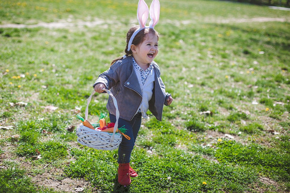A young girl with bunny ears headband cheerful and running outdoors with Easter basket filled with carrots, on a sunny grassy field.