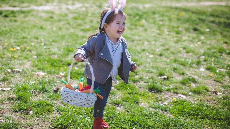 A young girl with bunny ears headband cheerful and running outdoors with Easter basket filled with carrots, on a sunny grassy field.