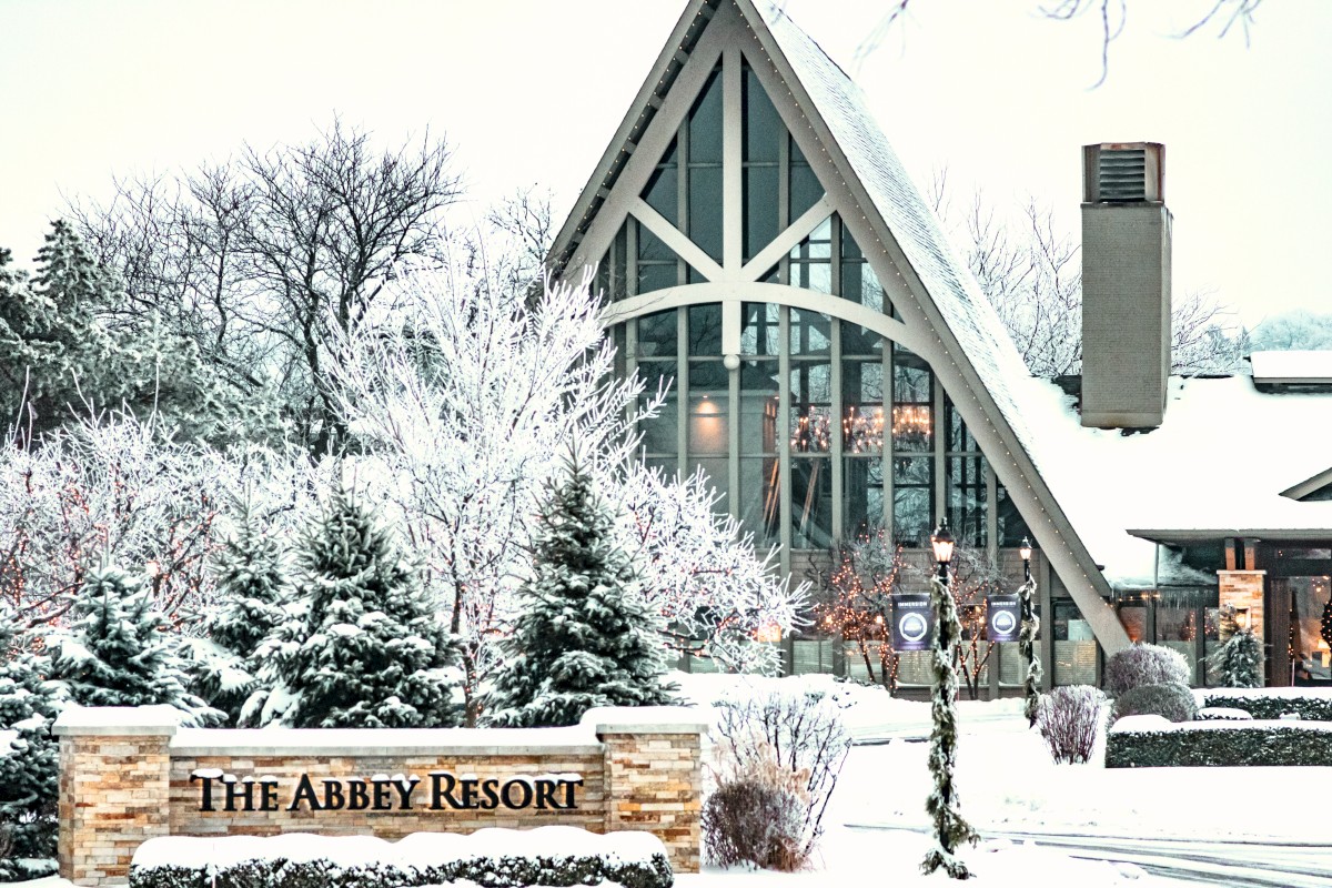 A snowy hotel entrance with a steep A-frame roof, frosted trees, and a sign reading “The Abbey Resort” in a wintery scene.