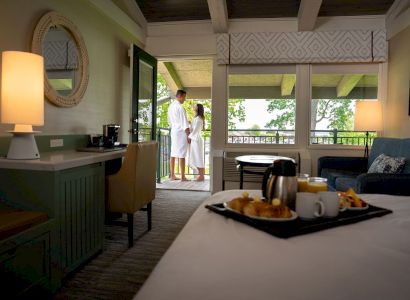 A cozy hotel room with a couple in bathrobes on the balcony, breakfast on the bed, and a warm lamp on a desk, ending the sentence.