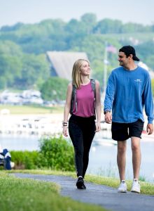 A couple is walking on a paved path beside a body of water, with a seated person in the background and greenery all around.