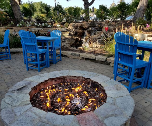 An outdoor seating area with blue chairs and tables surrounds a stone fire pit with a fire going, situated in a garden landscape.