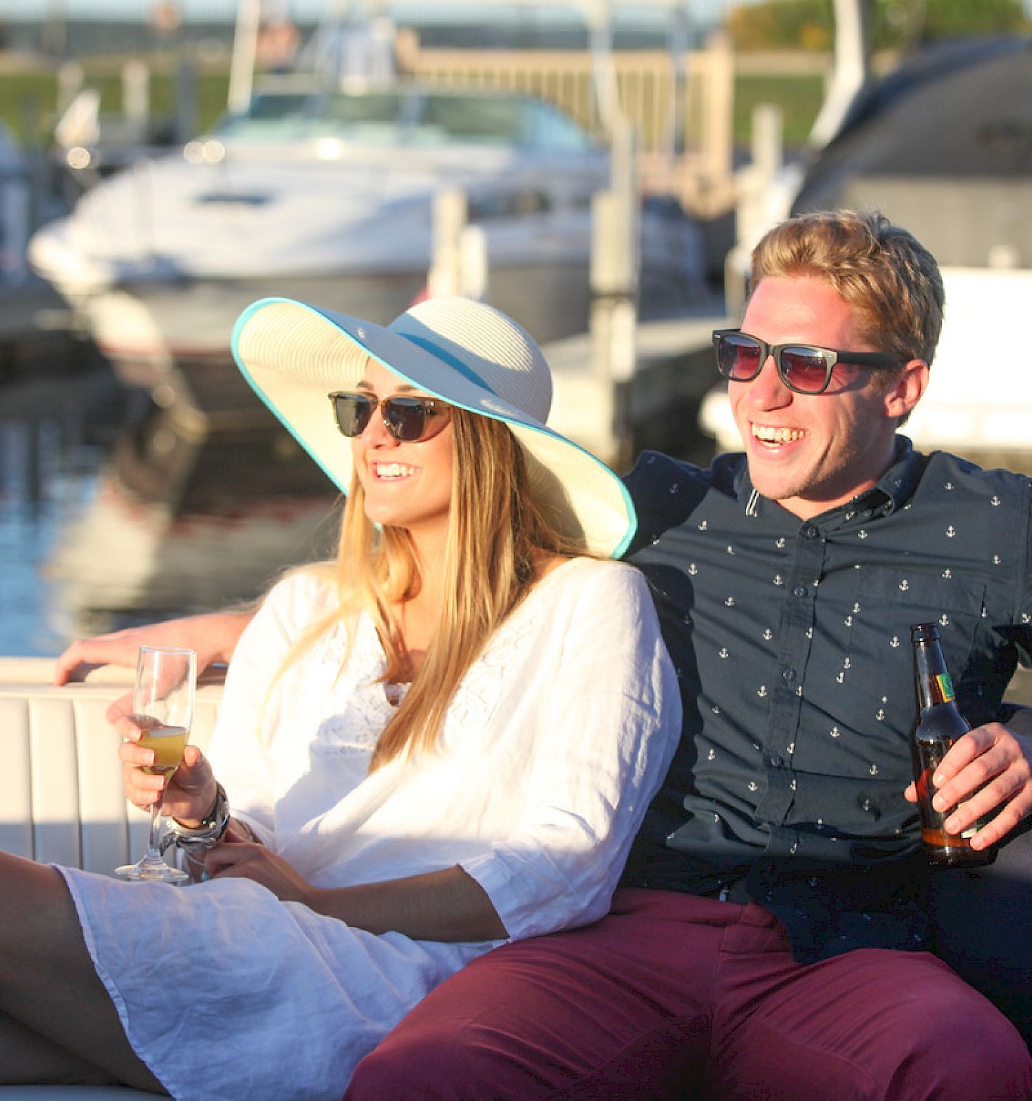 A man and woman are sitting on a boat, both wearing sunglasses, enjoying drinks, and smiling with boats and water in the background.