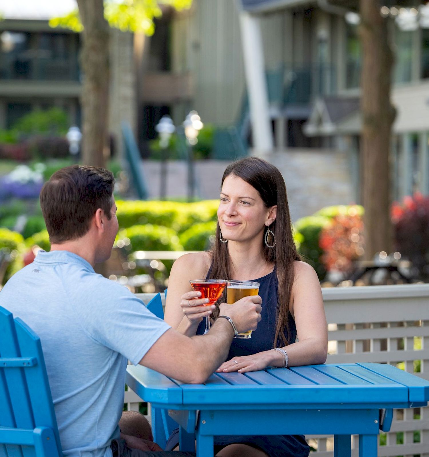 A man and woman sit at a blue table outdoors, clinking glasses in a cheerful setting with greenery and buildings in the background, smiling.