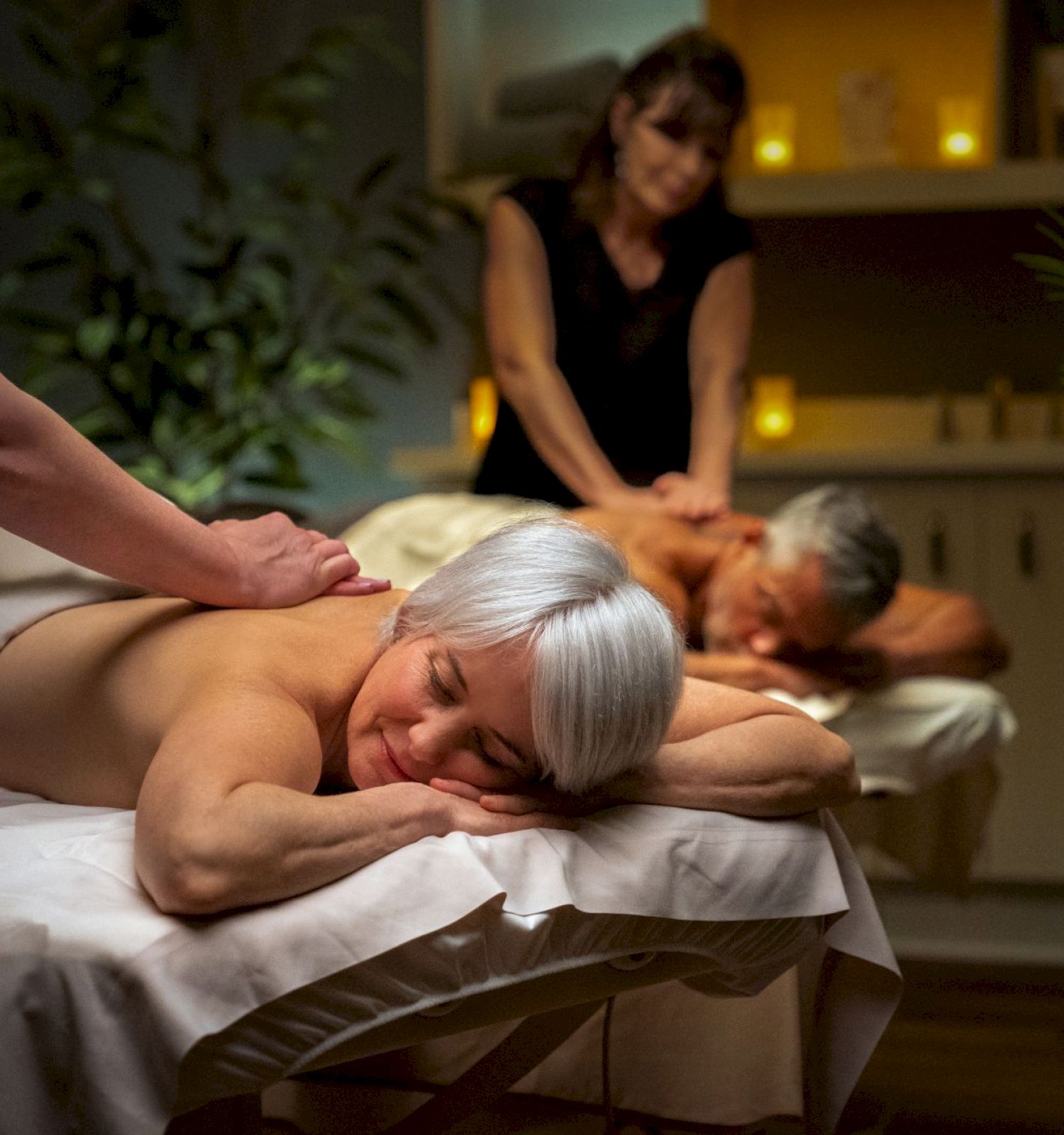 The image shows two people receiving massages on tables, attended by two masseuses in a dimly lit room with plants and candles.