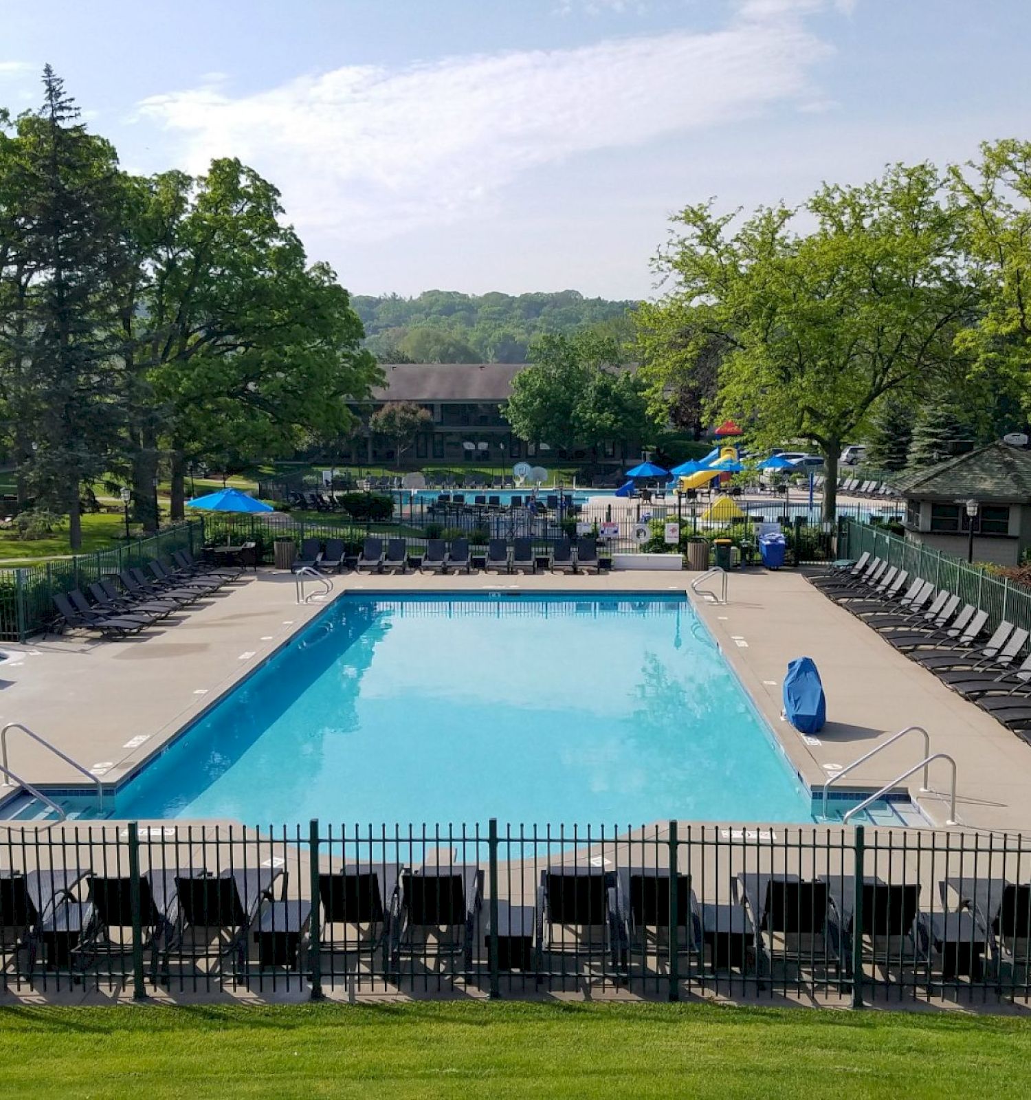 An outdoor swimming pool surrounded by lounge chairs and umbrellas, with trees and buildings in the background on a sunny day.