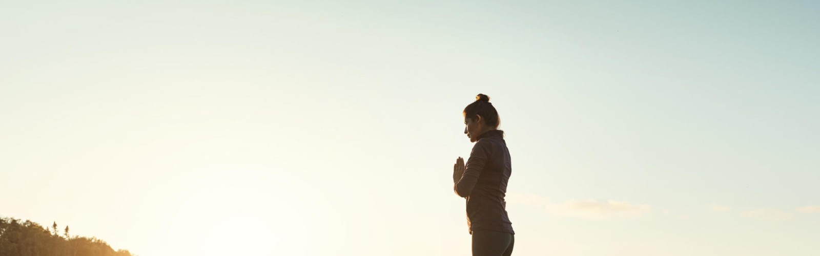 A person stands on a paddleboard on calm water during sunset, holding a yoga pose with hands in a prayer position, in a serene outdoor setting.