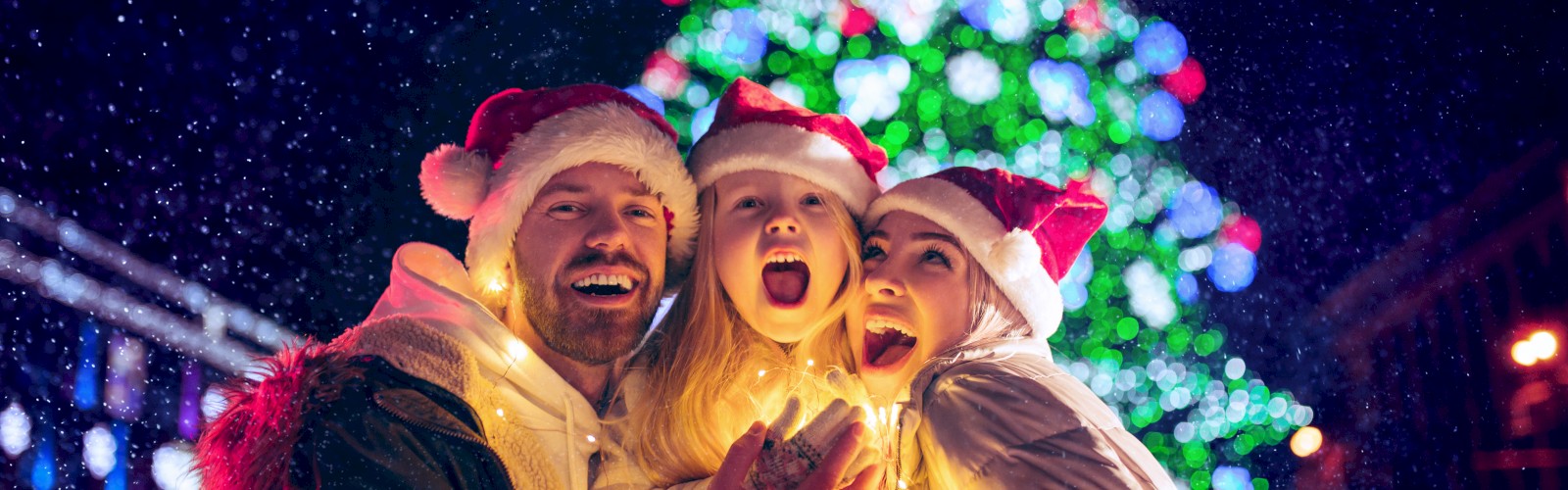A family wearing Santa hats is joyfully posing in front of a brightly lit Christmas tree at night, with festive decorations and lights in the background.