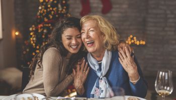 A young woman and an elderly woman are sharing a joyful moment at a festive dinner table with a Christmas tree and stockings in the background.