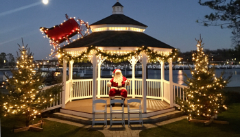 A gazebo is decorated with Christmas lights and garlands, featuring a Santa Claus figure and two Christmas trees at night.