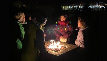 A group of children, wearing winter clothing, gather around a small fire pit at night while festive lights in the background illuminate the scene.