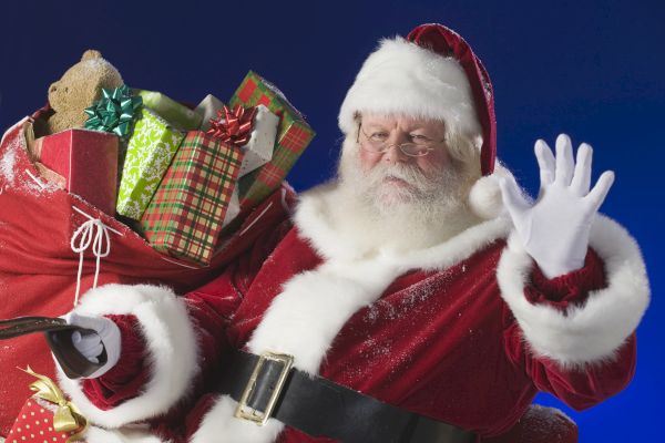 A person dressed as Santa Claus is holding a sack full of wrapped presents, waving with a gloved hand, against a blue background.