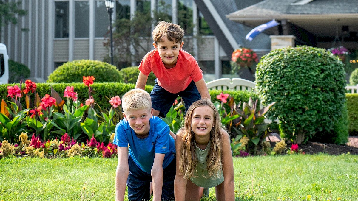 Three kids are forming a pyramid on a grassy lawn with flowers in the background, smiling and enjoying the outdoors.