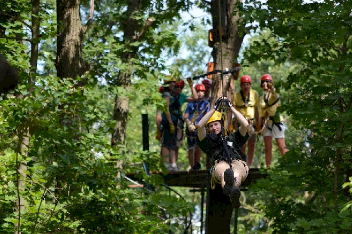 A person is zip-lining through a forest while a group waits on a platform in the background, all wearing helmets and harnesses.