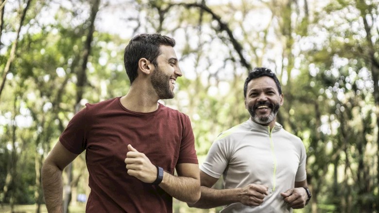 Two people are jogging and smiling in a wooded outdoor area, enjoying the fresh air and greenery around them.