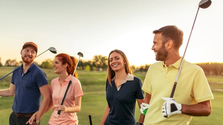 A group of four people smiling and holding golf clubs, walking on a golf course under a warm sunset.