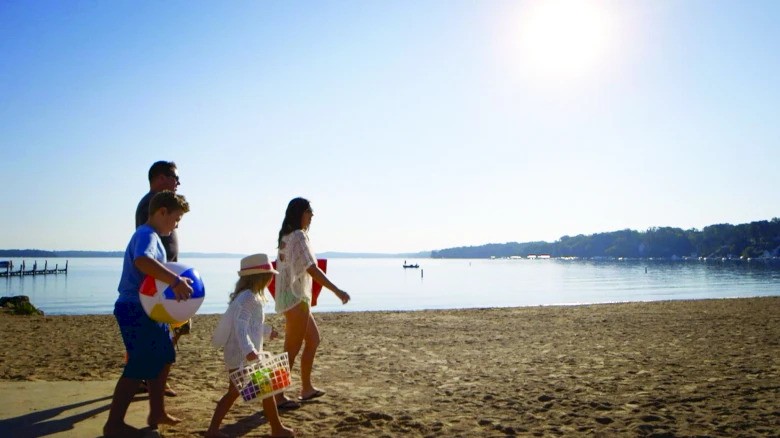 A family walks on a sunny beach, carrying beach balls and a cooler, with a calm sea and clear blue sky in the background.