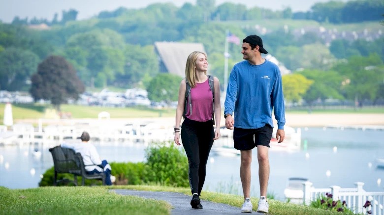 A couple is walking along a scenic lakeside path, surrounded by lush greenery, with a serene view and a person seated on a bench nearby.