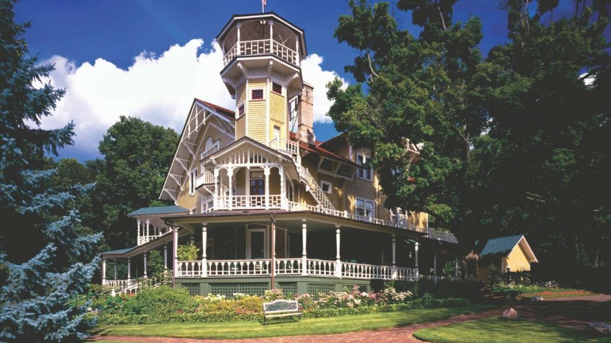 A multi-story Victorian-style house with ornate woodwork and a tower, surrounded by greenery and trees under a blue sky.