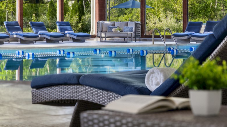 An indoor pool area with lounge chairs, a rolled towel, an open book, and a potted plant, surrounded by large windows and greenery.