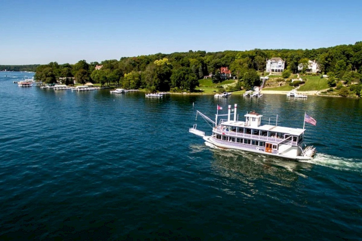 A boat is sailing on a lake near a lush, green shoreline with houses, under a clear blue sky.