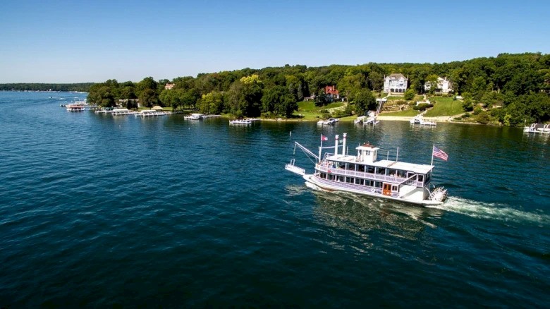 A boat is sailing on a lake near a lush, green shoreline with houses, under a clear blue sky.