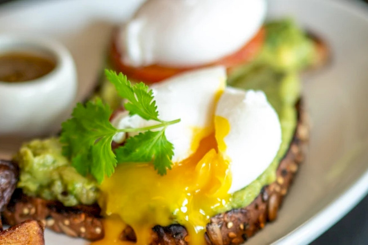 Avocado toast topped with a poached egg and garnished with cilantro, served with a side of sauce on a white plate.