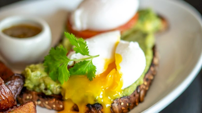Avocado toast topped with a poached egg and garnished with cilantro, served with a side of sauce on a white plate.