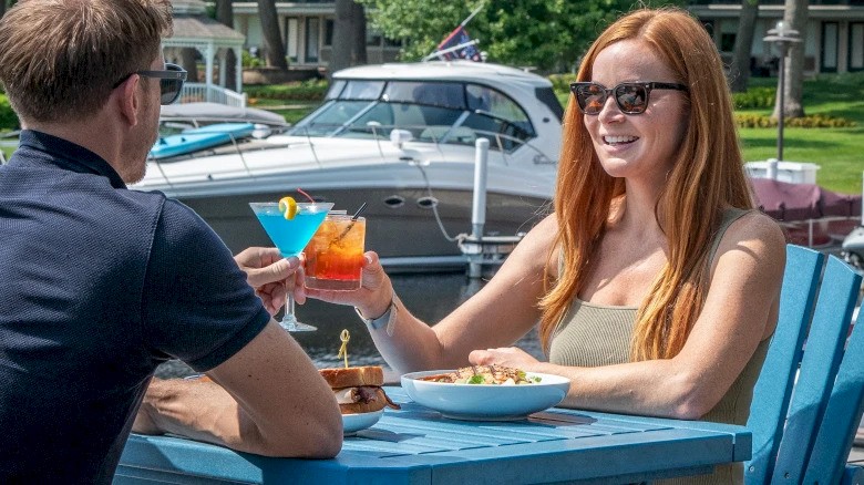 Two people are seated outdoors at a table by the water, enjoying drinks and food, with boats docked in the background.