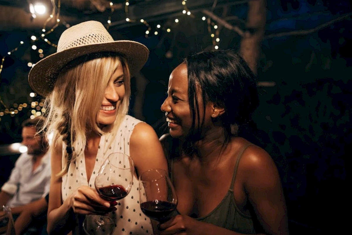 Two people are smiling at each other while holding wine glasses at an outdoor gathering with string lights in the background.