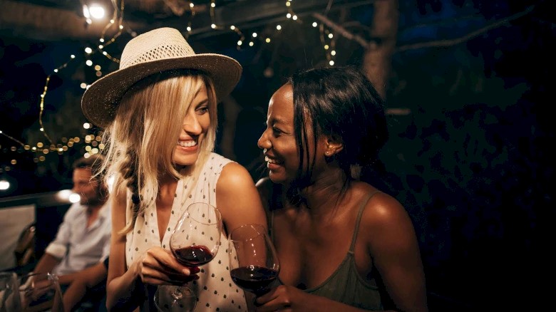Two people are smiling at each other while holding wine glasses at an outdoor gathering with string lights in the background.