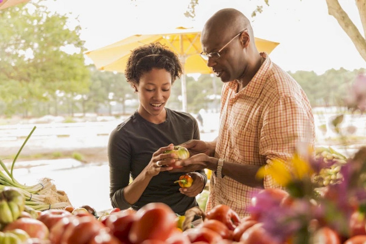 A couple is selecting fresh produce at an outdoor market, surrounded by a variety of colorful vegetables and flowers.