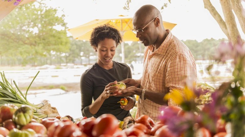 A couple is selecting fresh produce at an outdoor market, surrounded by a variety of colorful vegetables and flowers.