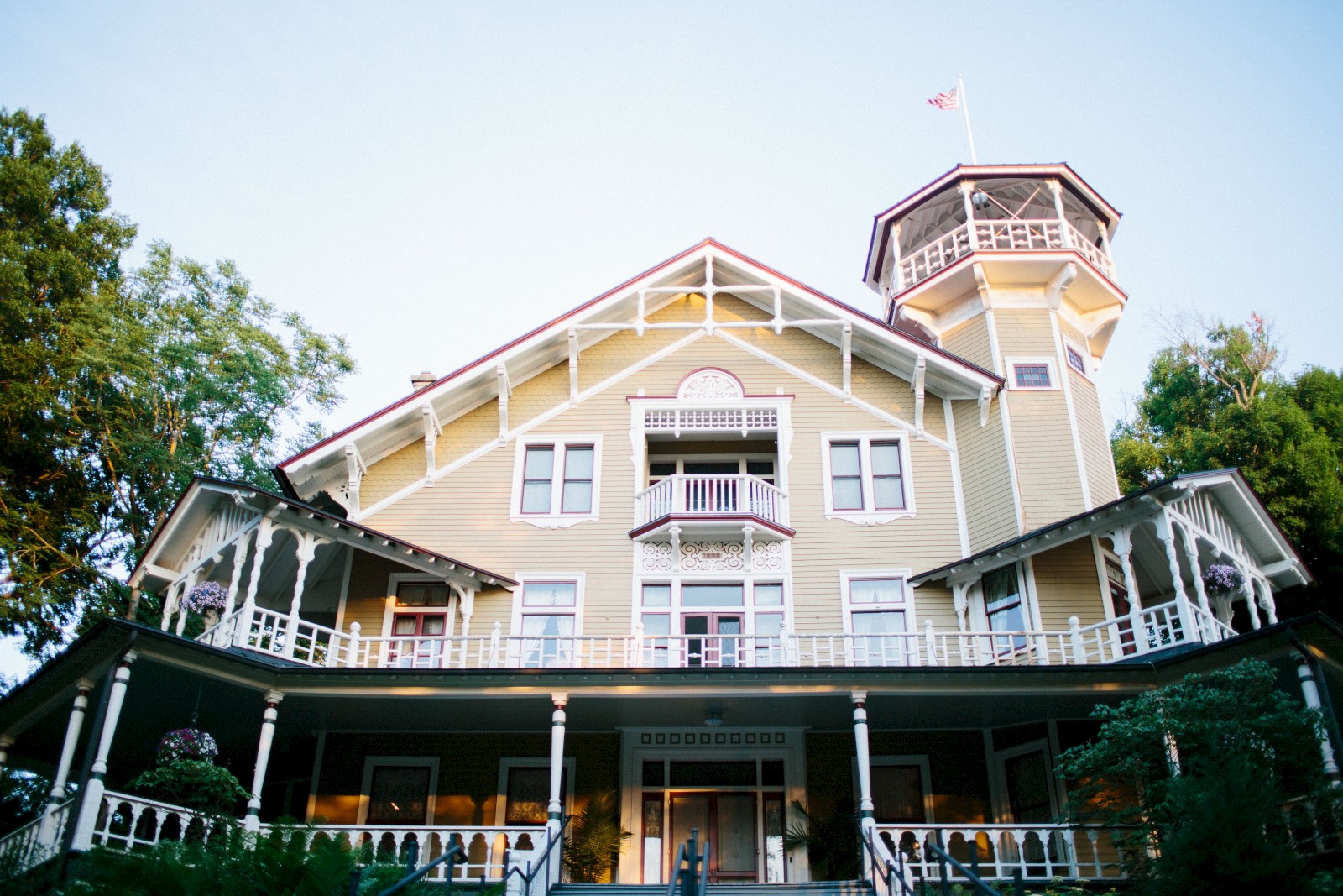 A large, historic-looking yellow house with a wraparound porch and a tower on the right, surrounded by trees under a clear sky.