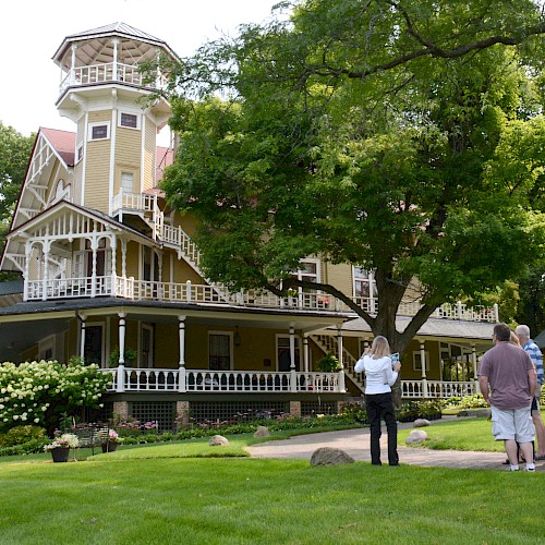 A group of people stands outside a large, ornate house with a wraparound porch and tower, surrounded by trees and greenery.
