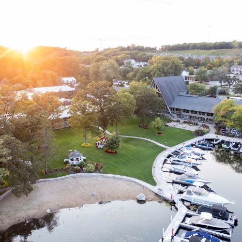 An aerial view of a lakeside marina with boats, a sandy beach, green lawns, trees, and buildings under a setting sun.