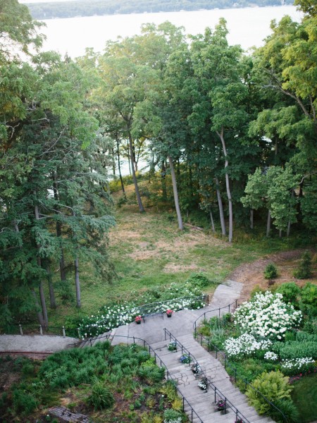 A garden with pathways surrounded by greenery and flowers, leading to a wooded area near a body of water.
