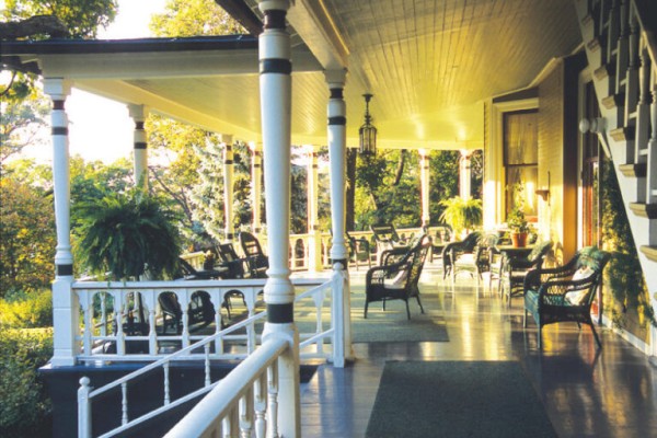 A spacious porch with wicker furniture, potted plants, and a wooden railing, bathed in sunlight, overlooks lush greenery.