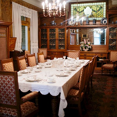 A vintage dining room with a long table set for a meal, surrounded by ornate wooden furniture and a chandelier.