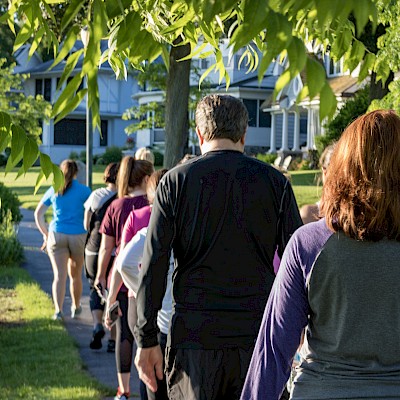 A group of people is walking on a sidewalk in a residential area, surrounded by greenery and houses.