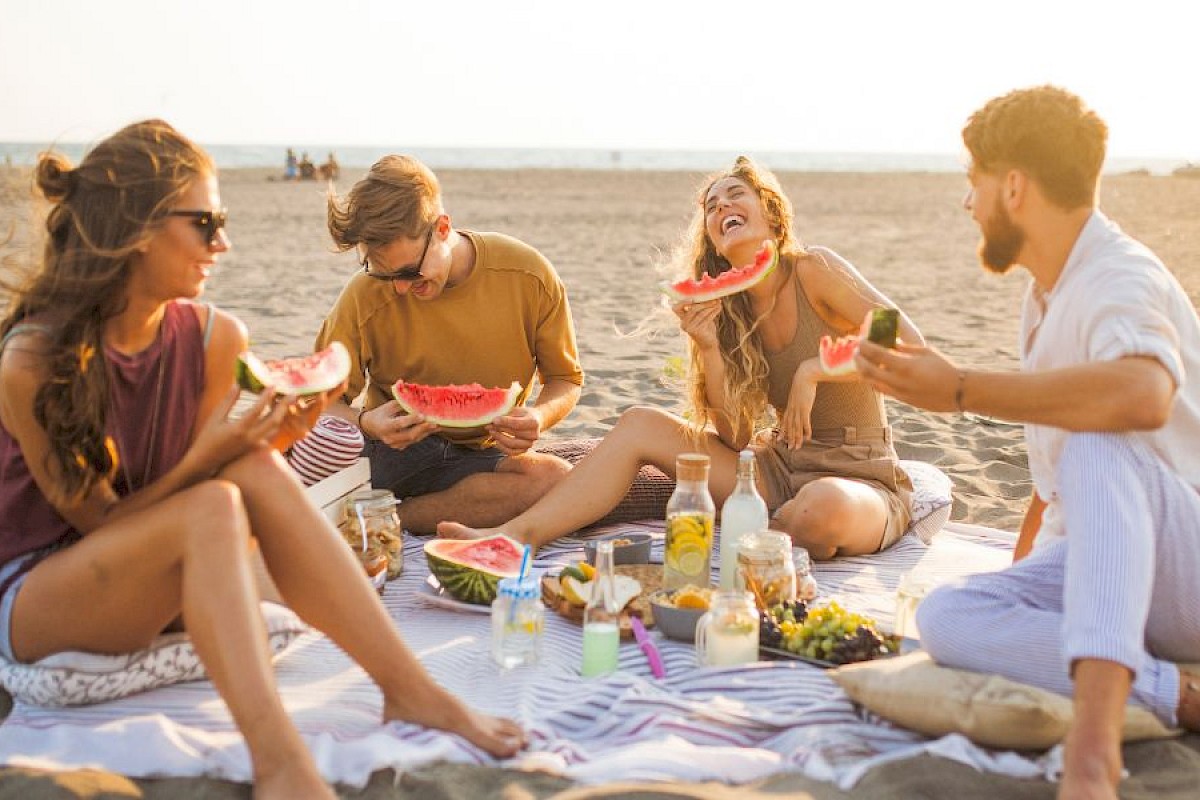A group of four friends enjoying watermelon and drinks during a sunny beach picnic on the sand.