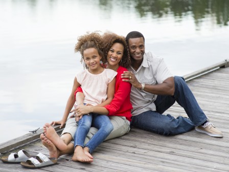 A joyful family of three sits on a wooden dock beside a calm lake, exuding warm vibes and smiles in a peaceful, scenic setting.