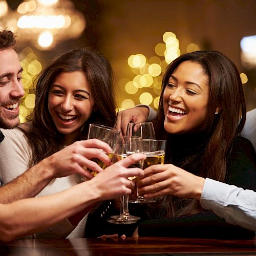 A group of five people is smiling and clinking their drinks together in a festive, warmly lit setting, likely celebrating or socializing.