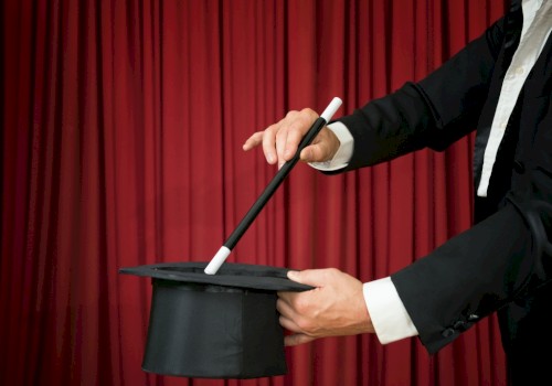 A magician is holding a top hat and a wand against a red curtain backdrop, suggesting a magic trick about to be performed.