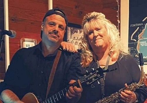 Two people pose with musical instruments, one holding a guitar and the other a saxophone, smiling indoors near a lit sign and wooden background.