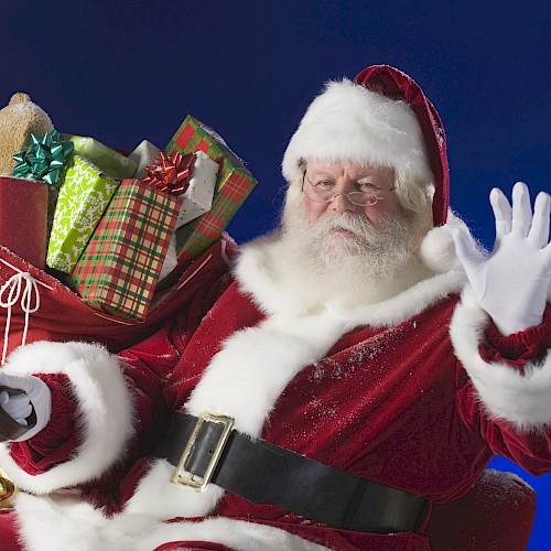 A person dressed as Santa Claus is waving, with a large red sack filled with wrapped gifts and a teddy bear over their shoulder, against a blue background.